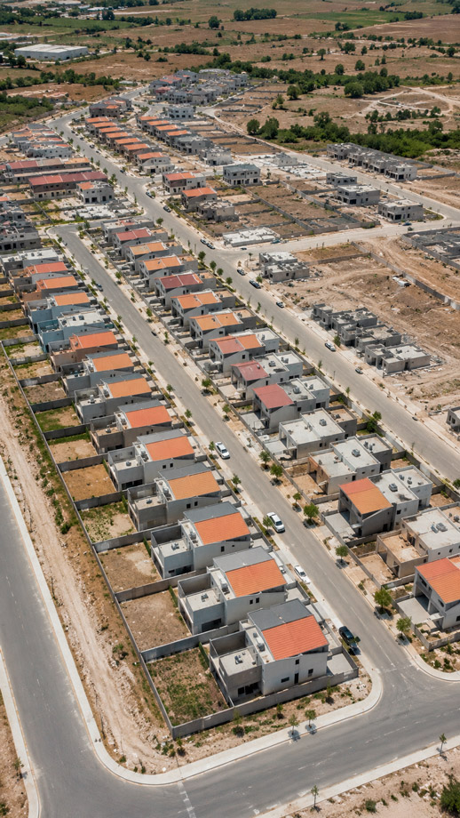 Fotografía aérea de dron de un fraccionamiento residencial en construcción y parcialmente terminado, hileras de casas con techos similares en tonos grises y terracota, calles pavimentadas recién trazadas, algunos lotes aún en etapa de cimentación visibles, vegetación incipiente, luz de día clara, composición geométrica ordenada vista desde arriba, paleta grafito y tierra, sin texto ni logotipos.